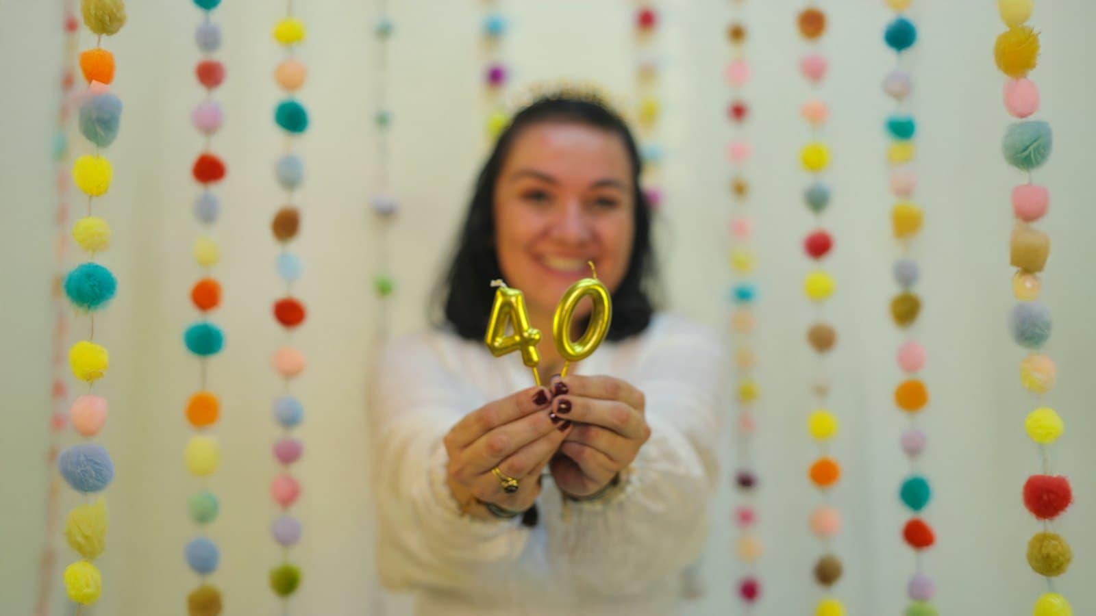 Smiling woman holding gold number 40 birthday candles in front of colourful pom-pom garlands