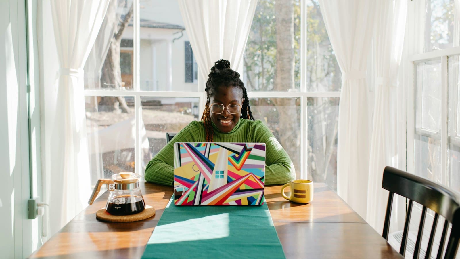 A woman sits at a sunny table at home checking her application on a laptop