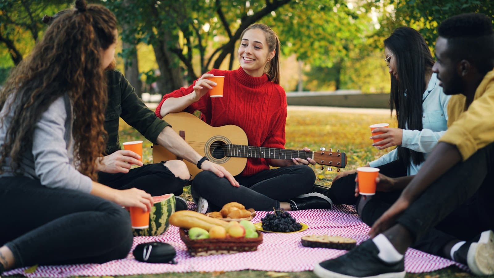 Group of friends sharing a relaxed picnic in a park with a guitar and drinks