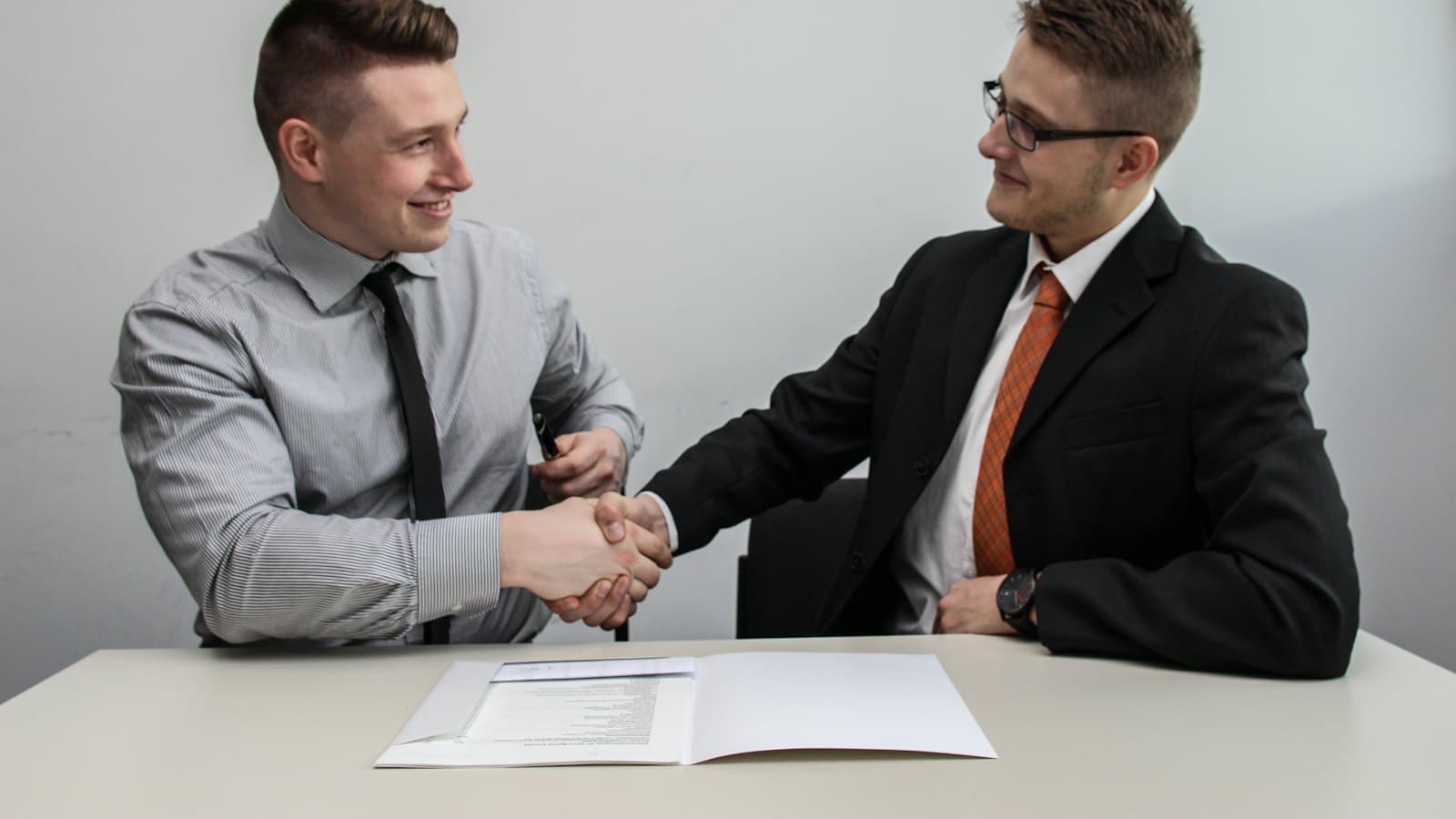 Two smiling men shaking hands across a desk over an employment contract