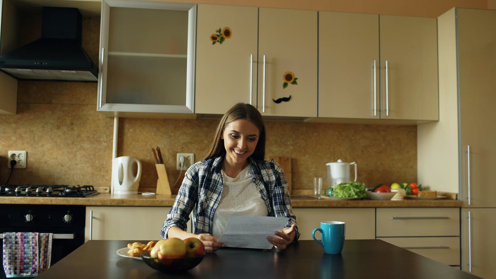 A smiling woman reading a letter at her kitchen table with coffee and fruit