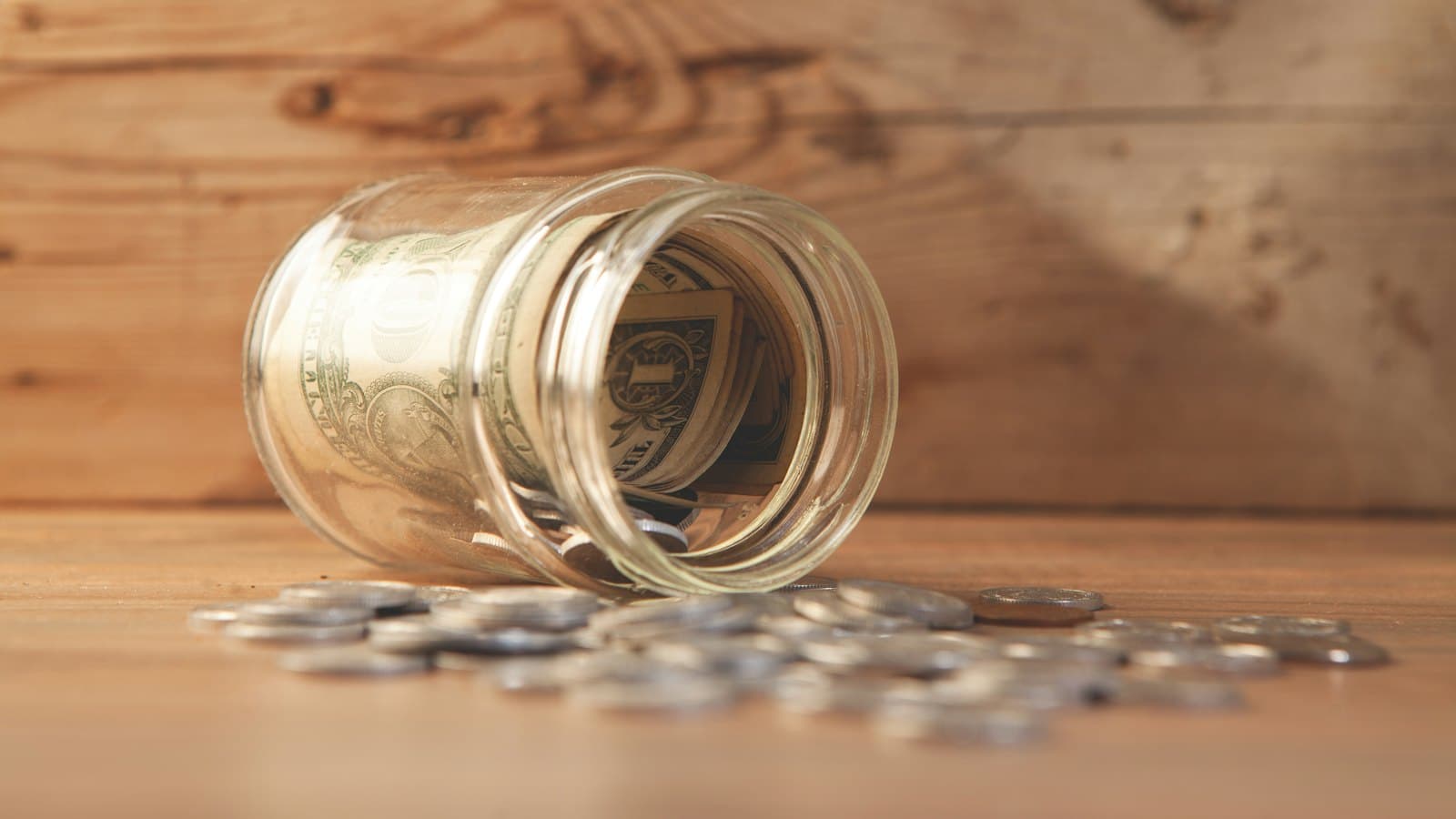 A glass savings jar with banknotes and coins spilling onto a table