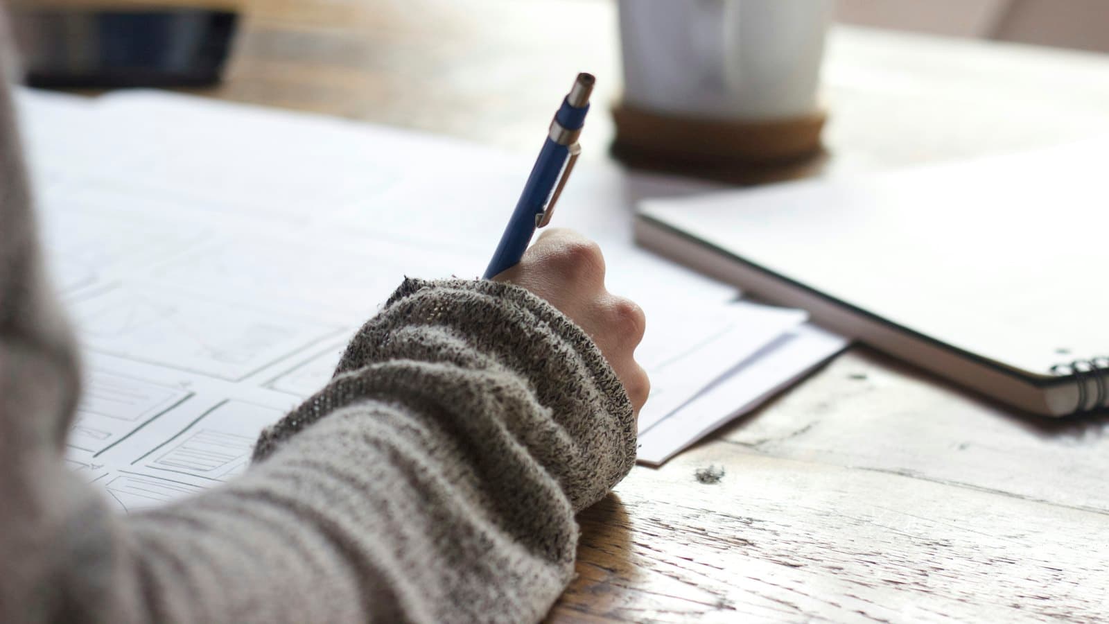 A person writing study notes at a wooden table beside a white mug