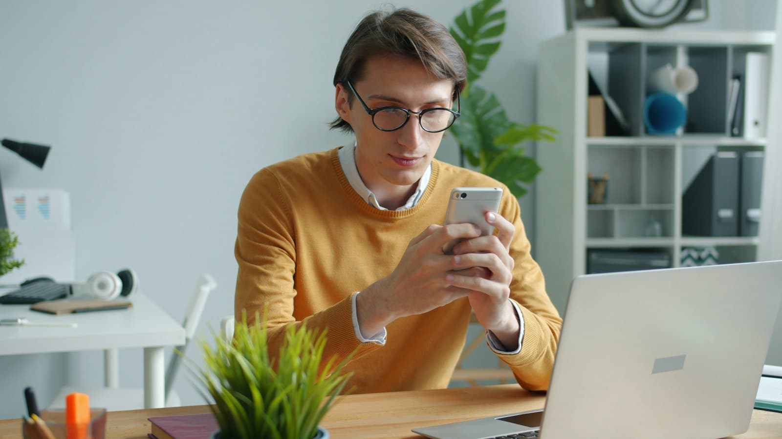 Man in glasses carefully reading a message on his phone at a desk beside a laptop