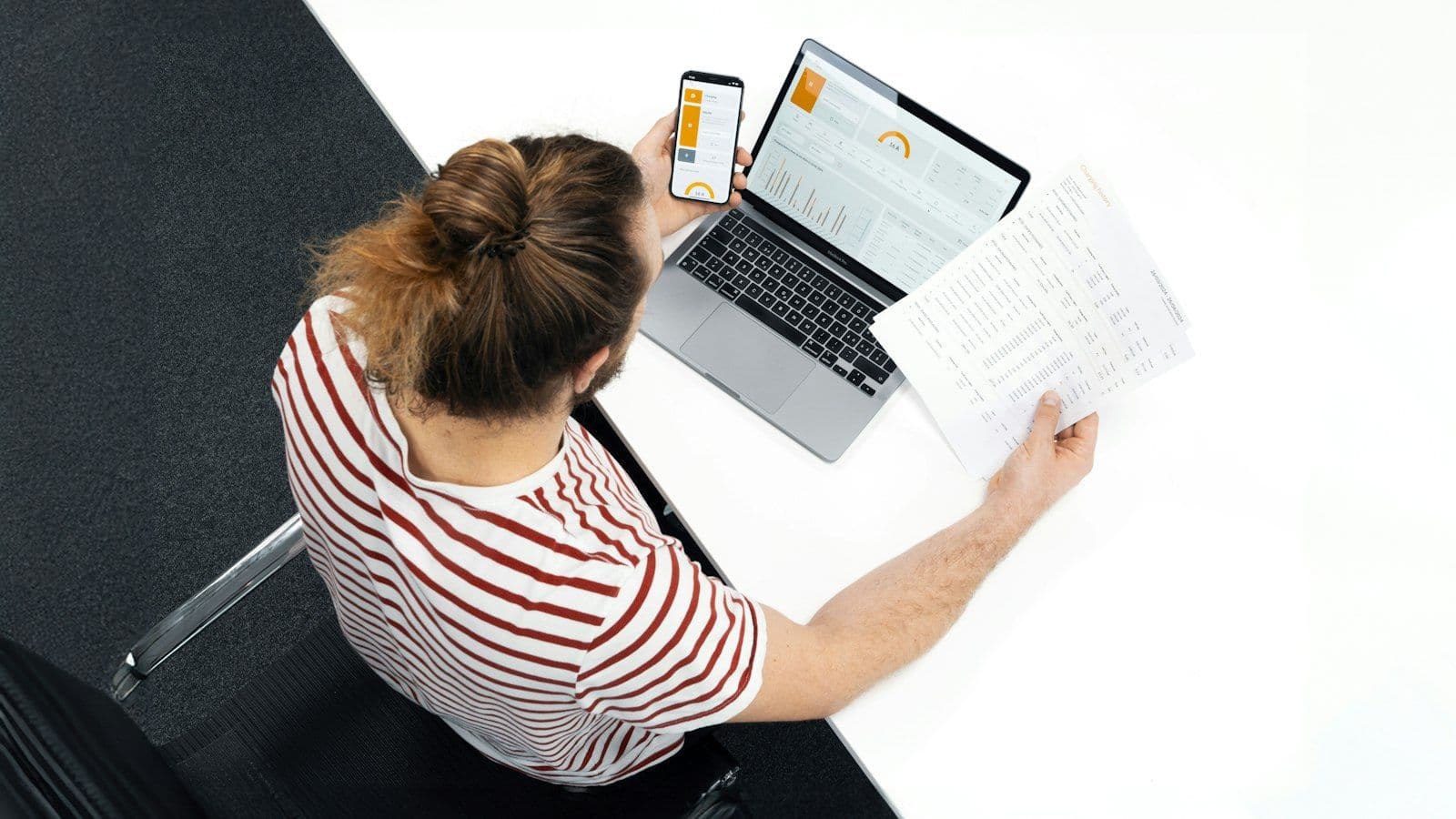 A person at a desk checking a printed document against a laptop and a phone