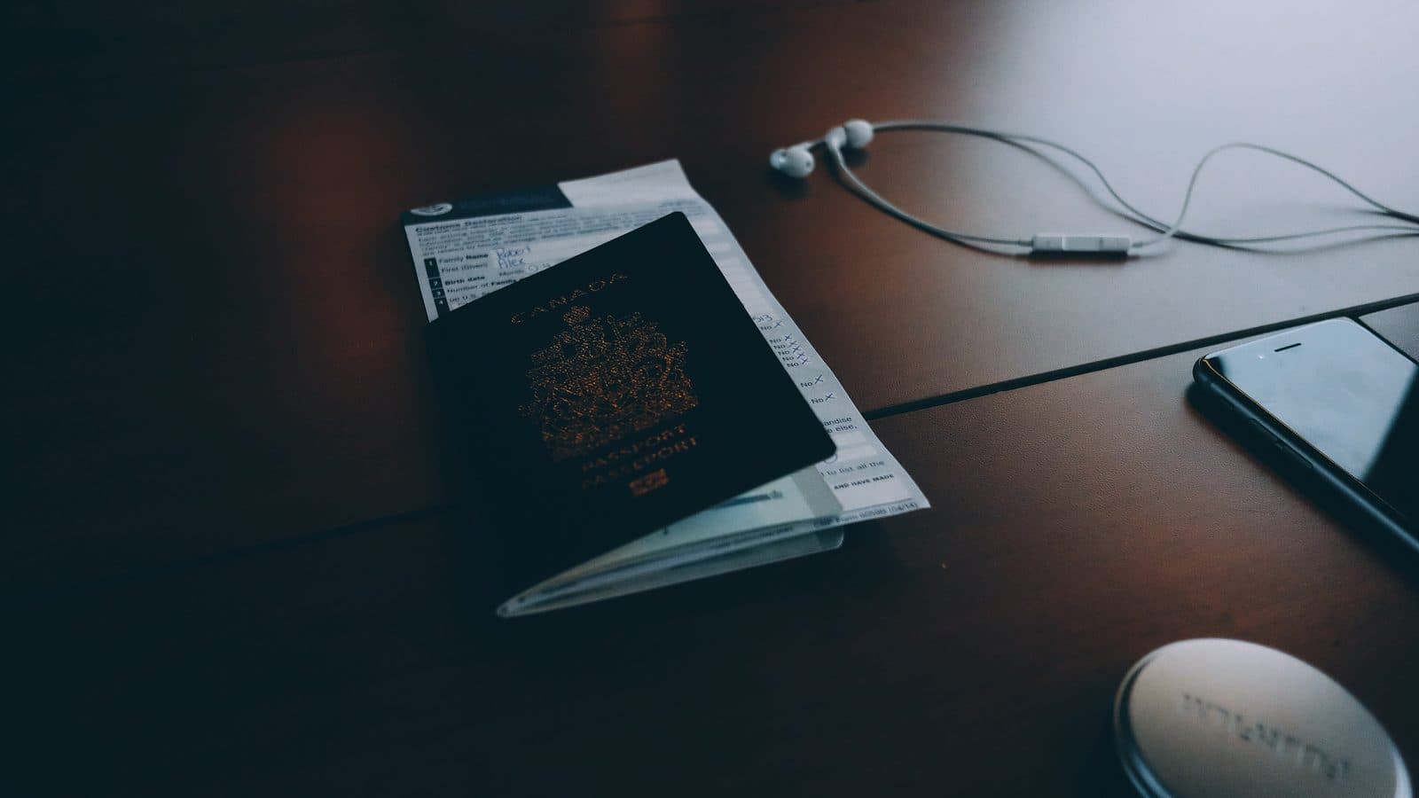 A Canadian passport resting on a customs declaration form on a dark wooden desk beside earphones and a phone