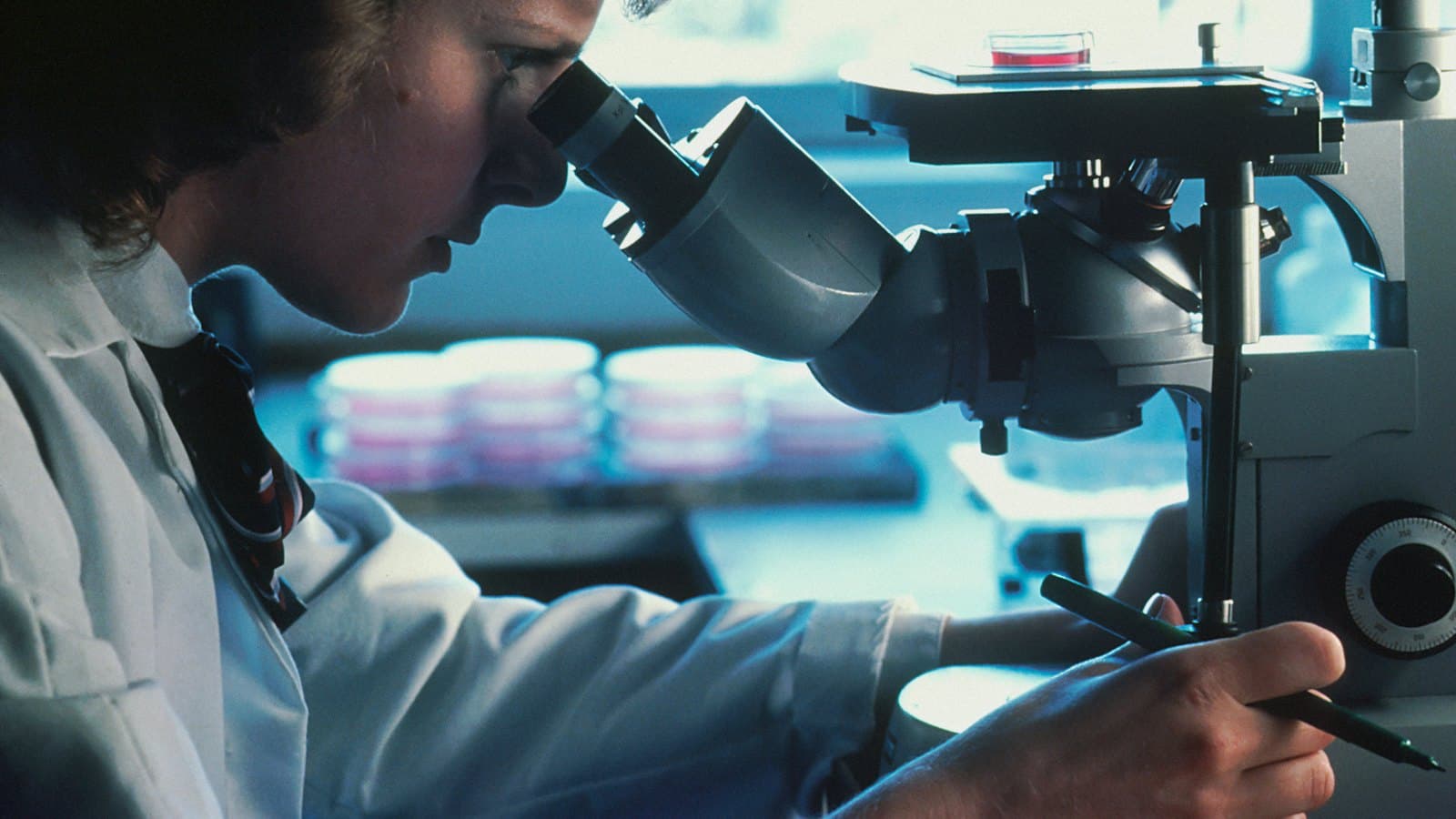 A scientist in a white lab coat looking through a microscope in a laboratory