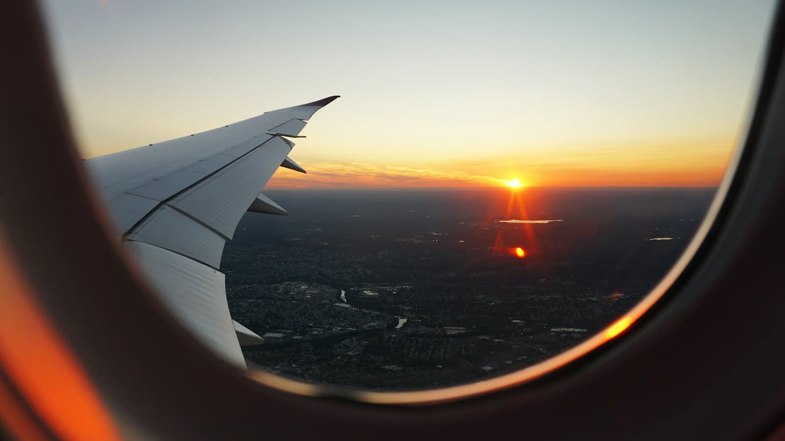 An airplane wing seen through the cabin window as the sun sets over a city