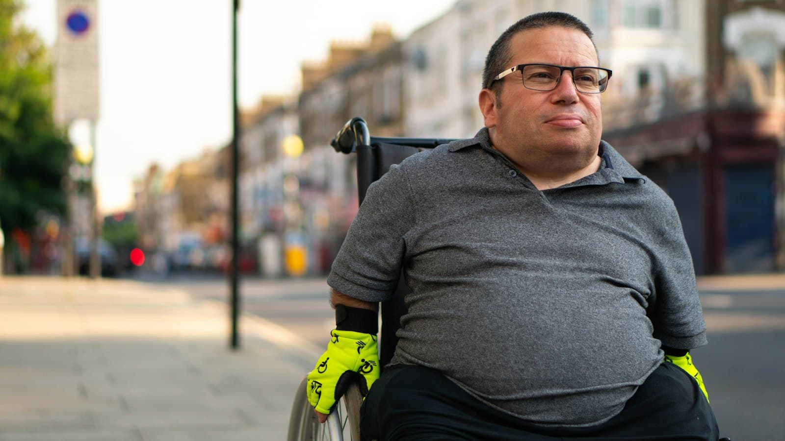 A man in glasses sits calmly in his wheelchair on a quiet, sunny city street