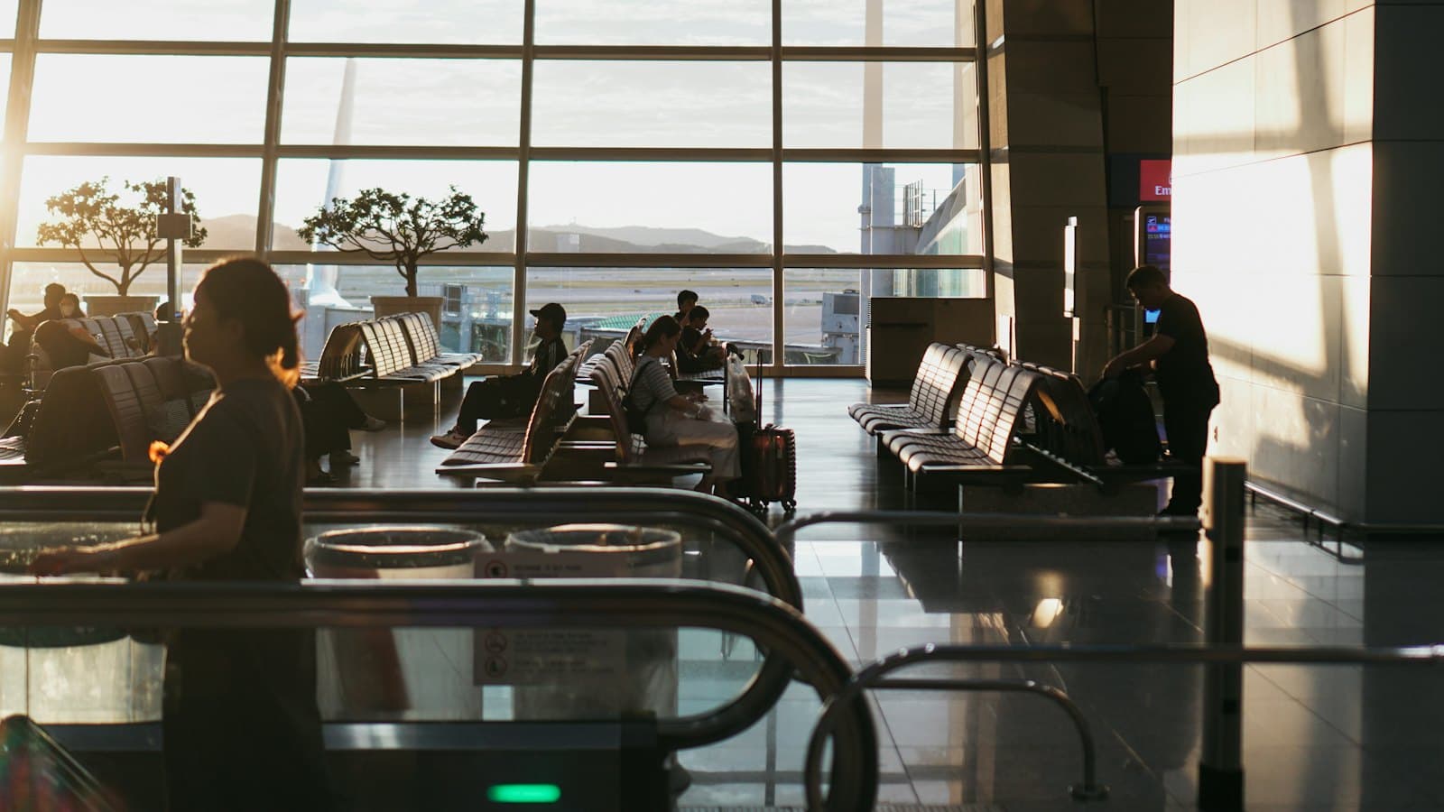 Travellers wait on benches in a quiet, sunlit airport departure lounge at sunset
