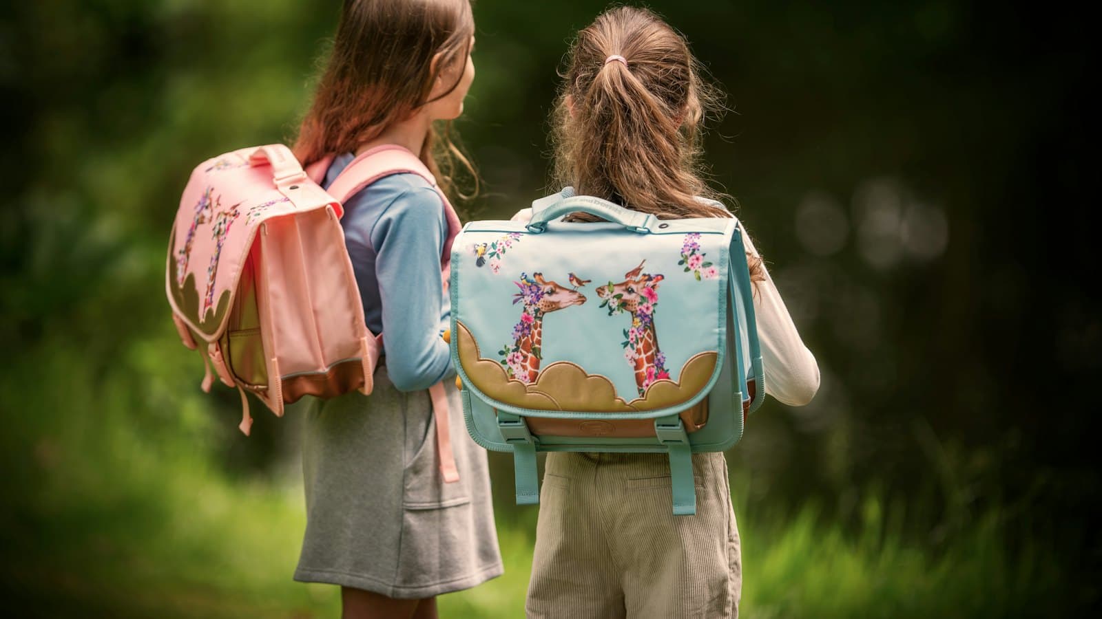 Two young girls with colourful school backpacks, seen from behind in a green park