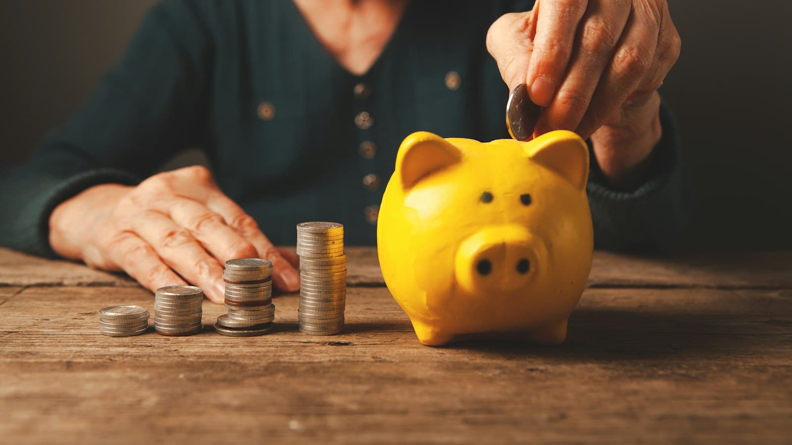 Older woman adding a coin to a yellow piggy bank beside stacks of coins