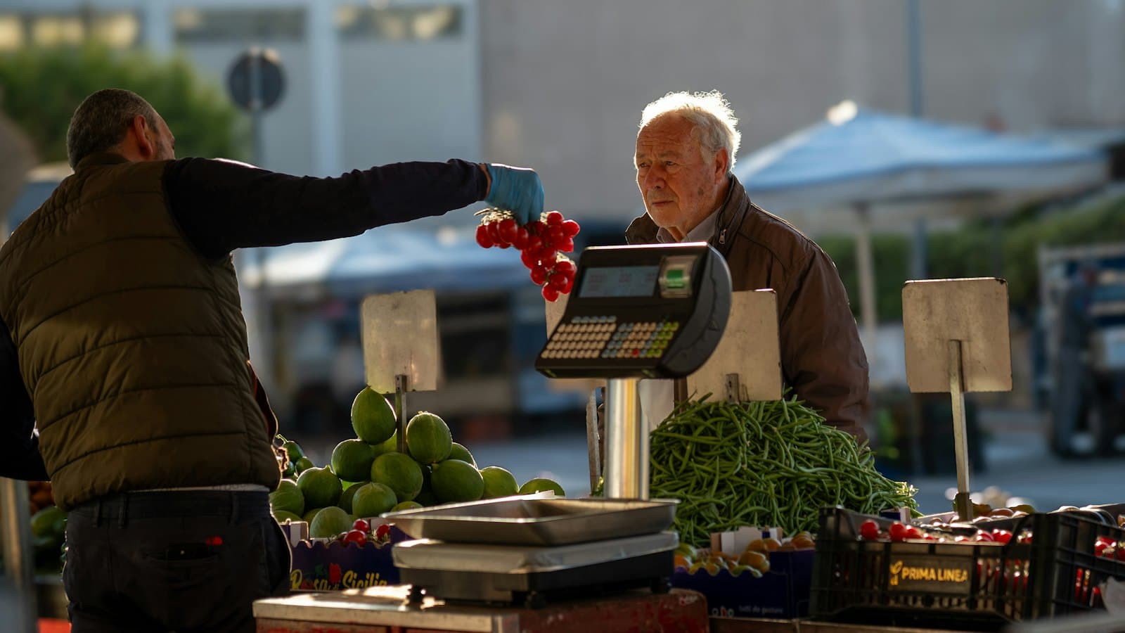 A market vendor handing cherry tomatoes across a weighing scale to an older customer