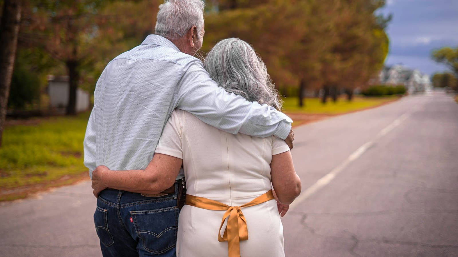 An older couple with grey hair walking arm in arm down a quiet tree-lined road