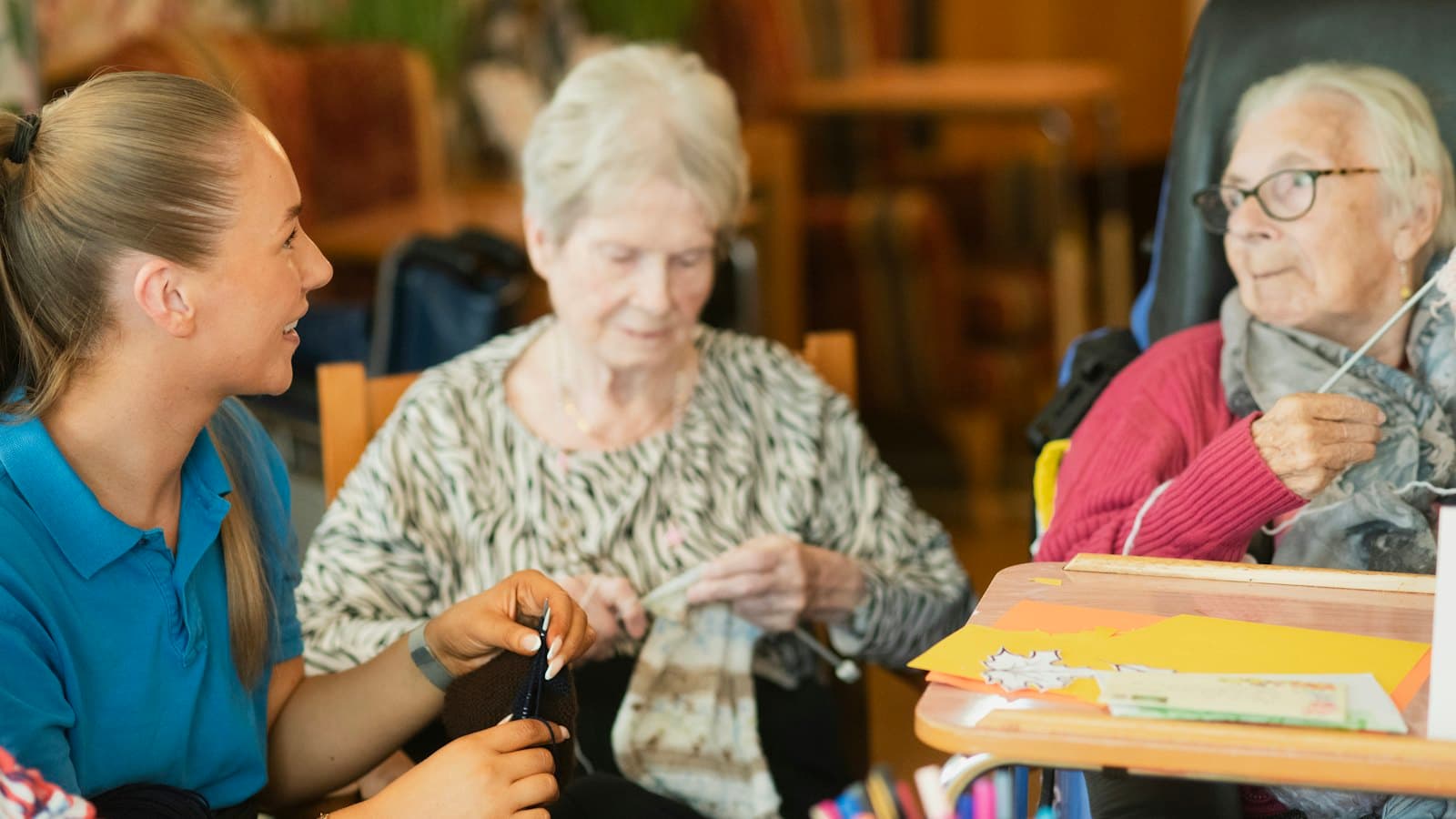 Smiling care worker knitting alongside two older women in a care home