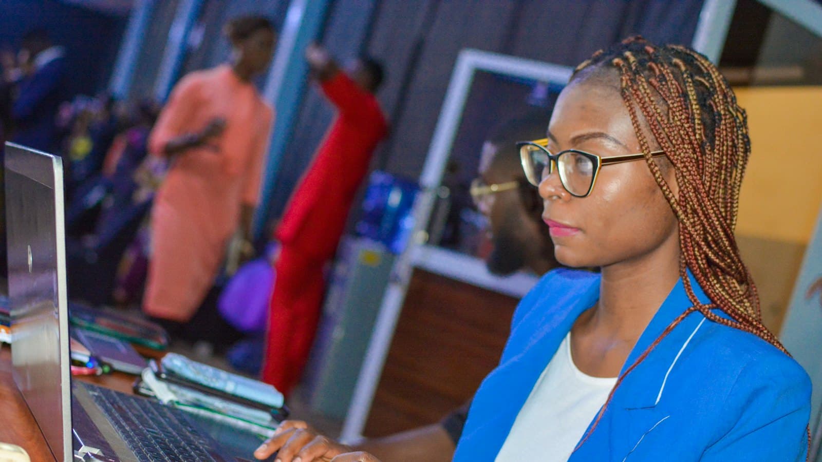 Woman in glasses and a blue blazer working on a laptop in a busy office