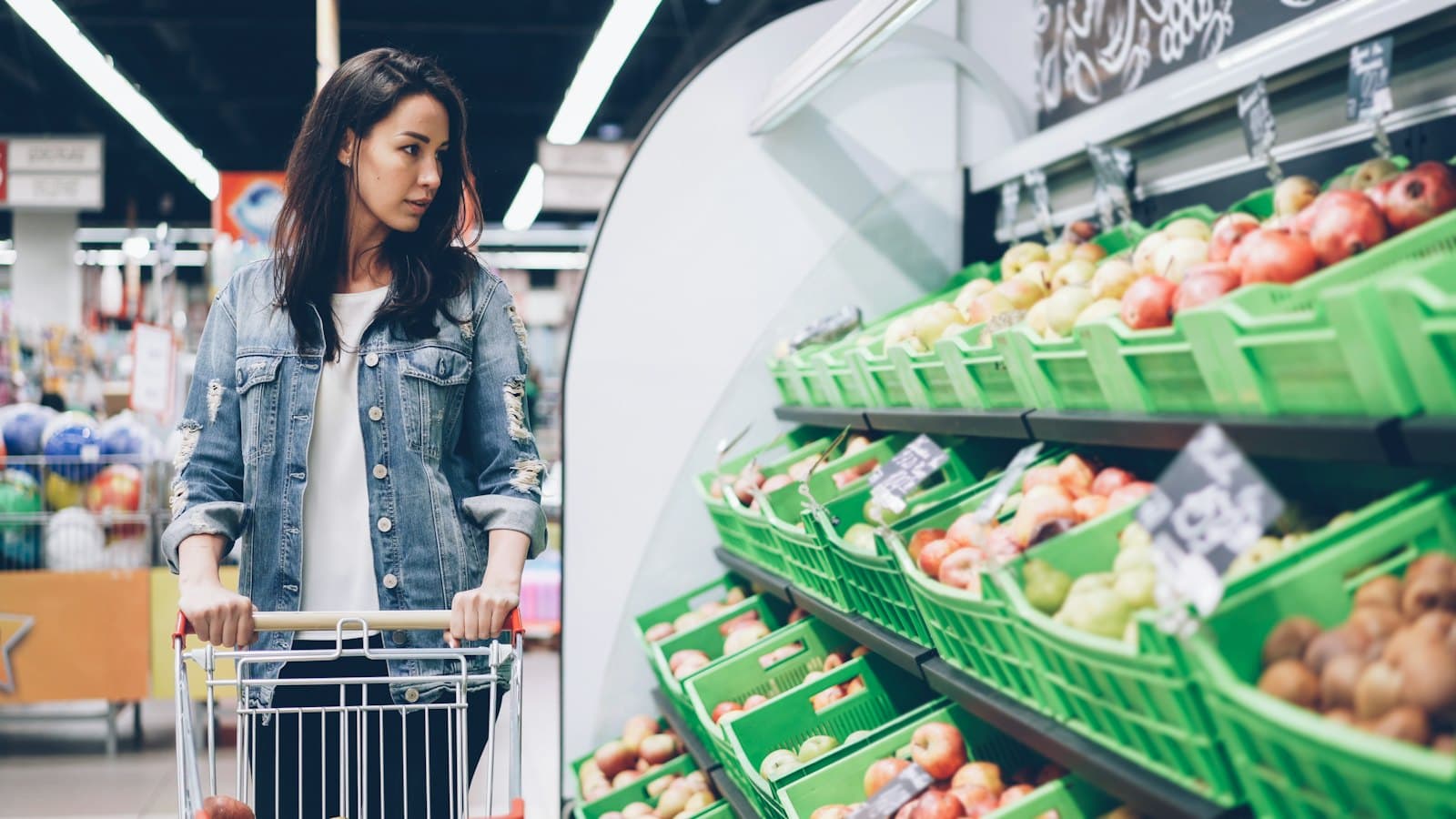 A woman pushing a shopping cart past fruit shelves in a supermarket
