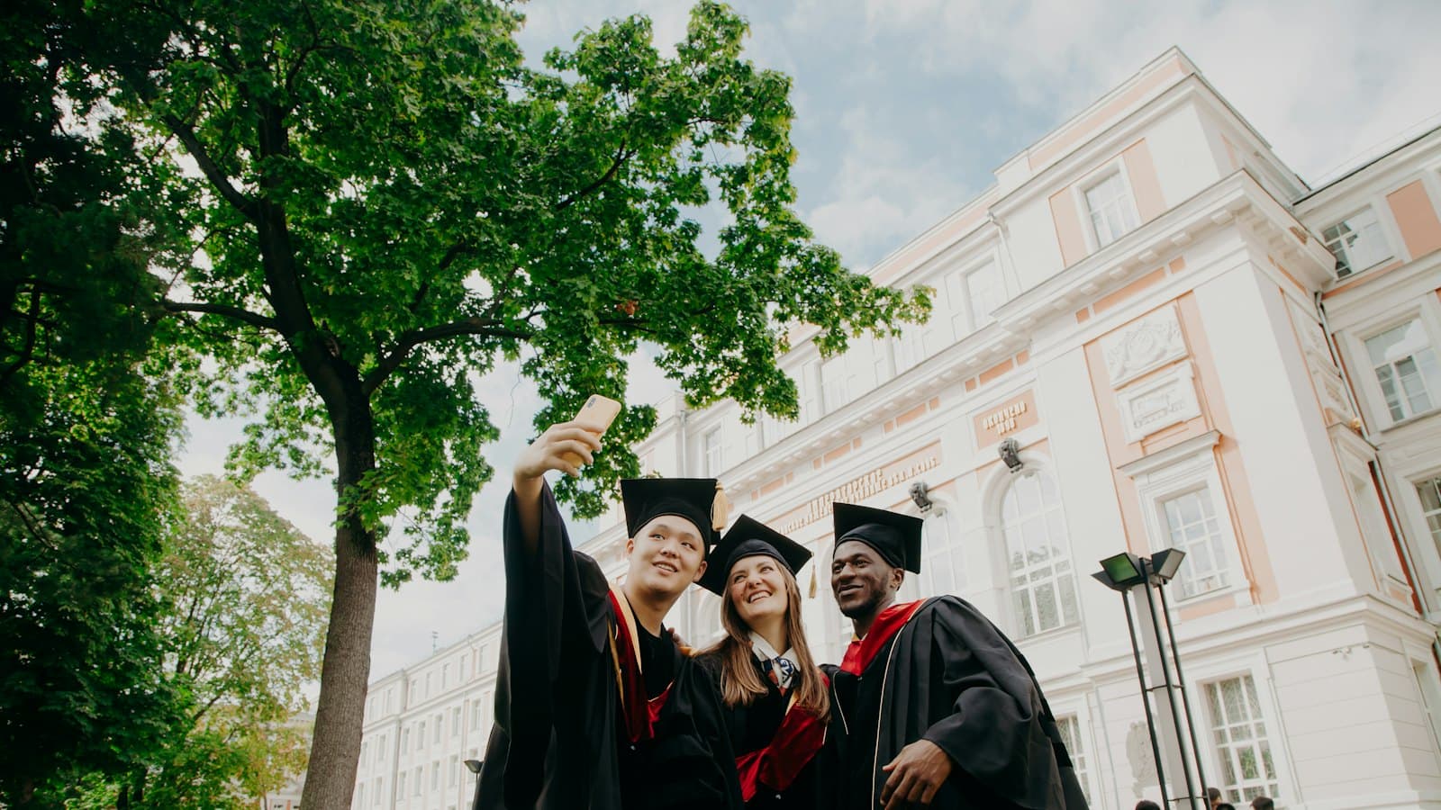 Three graduates in caps and gowns taking a selfie outside a university building