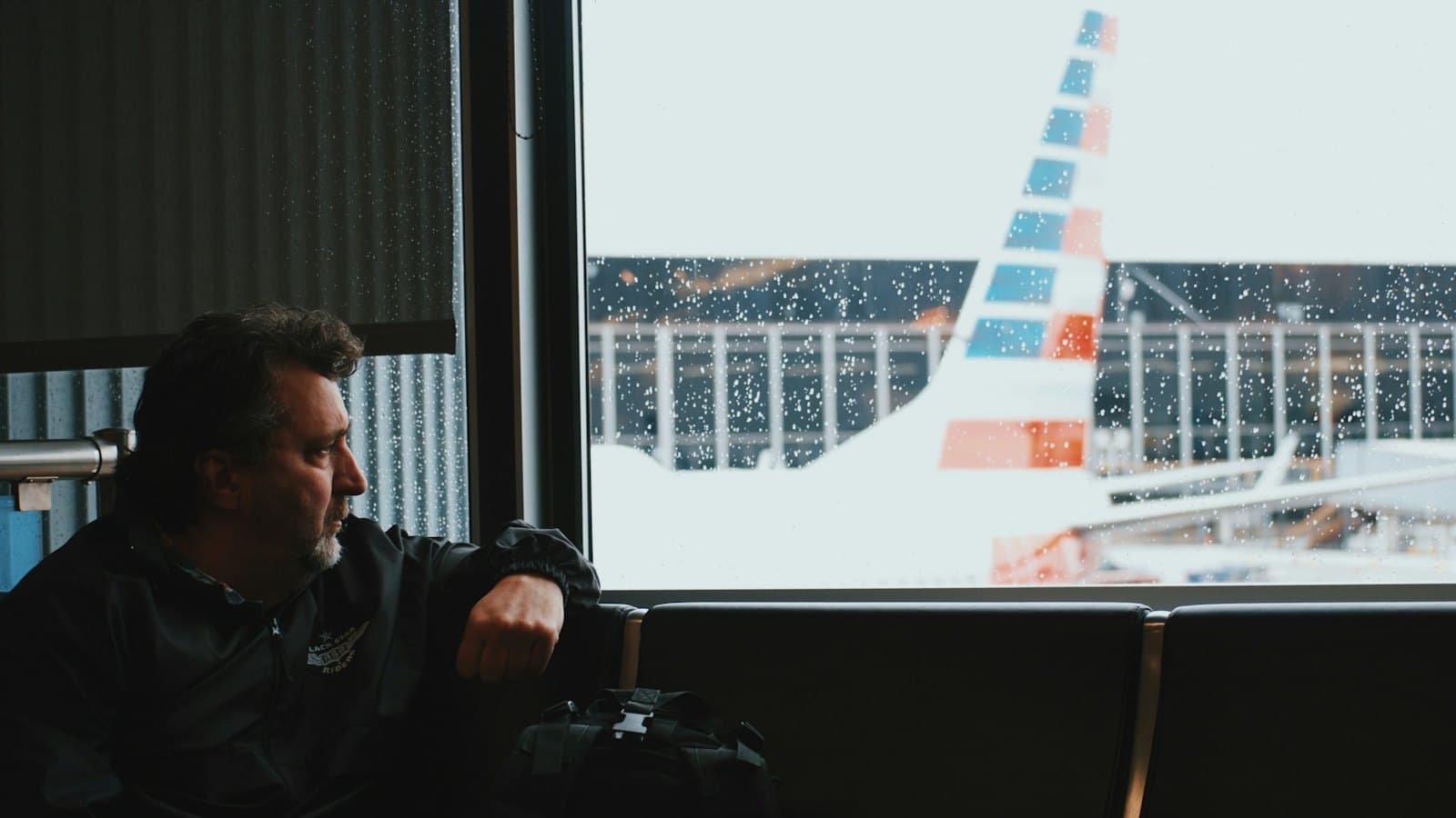 A middle-aged man at an airport gate looking out at a parked plane on a rainy day