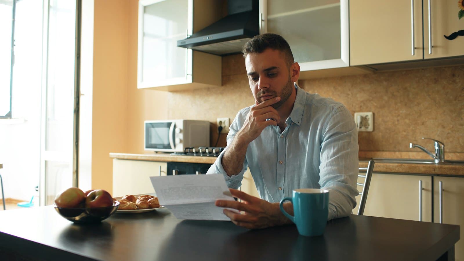 Man at a kitchen table thoughtfully reading a letter beside a cup of tea