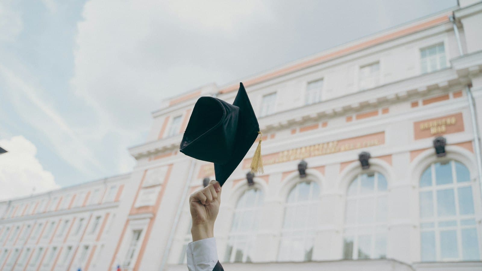 A hand holding a black graduation cap aloft in front of a large university building