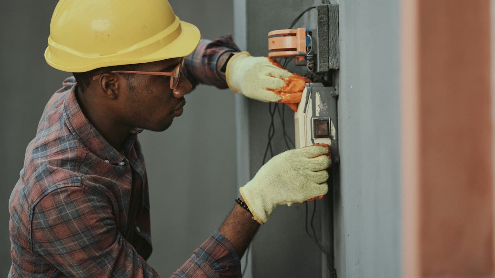 Electrician in a yellow hard hat and work gloves fitting a wall-mounted switch box