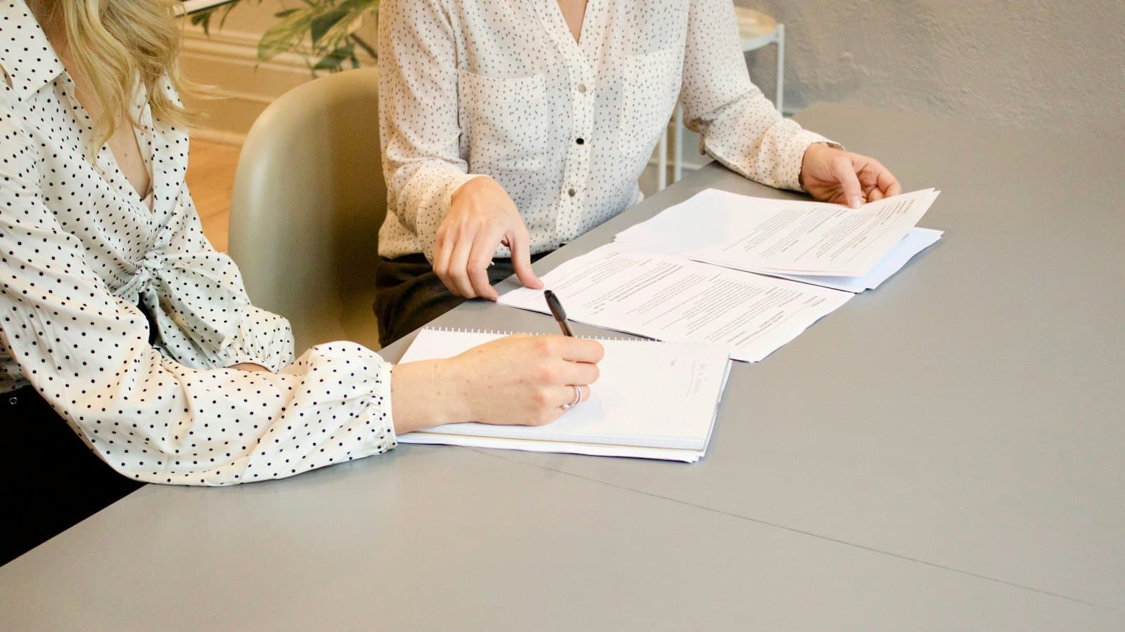 Woman taking notes while another woman points at printed contract pages on a table