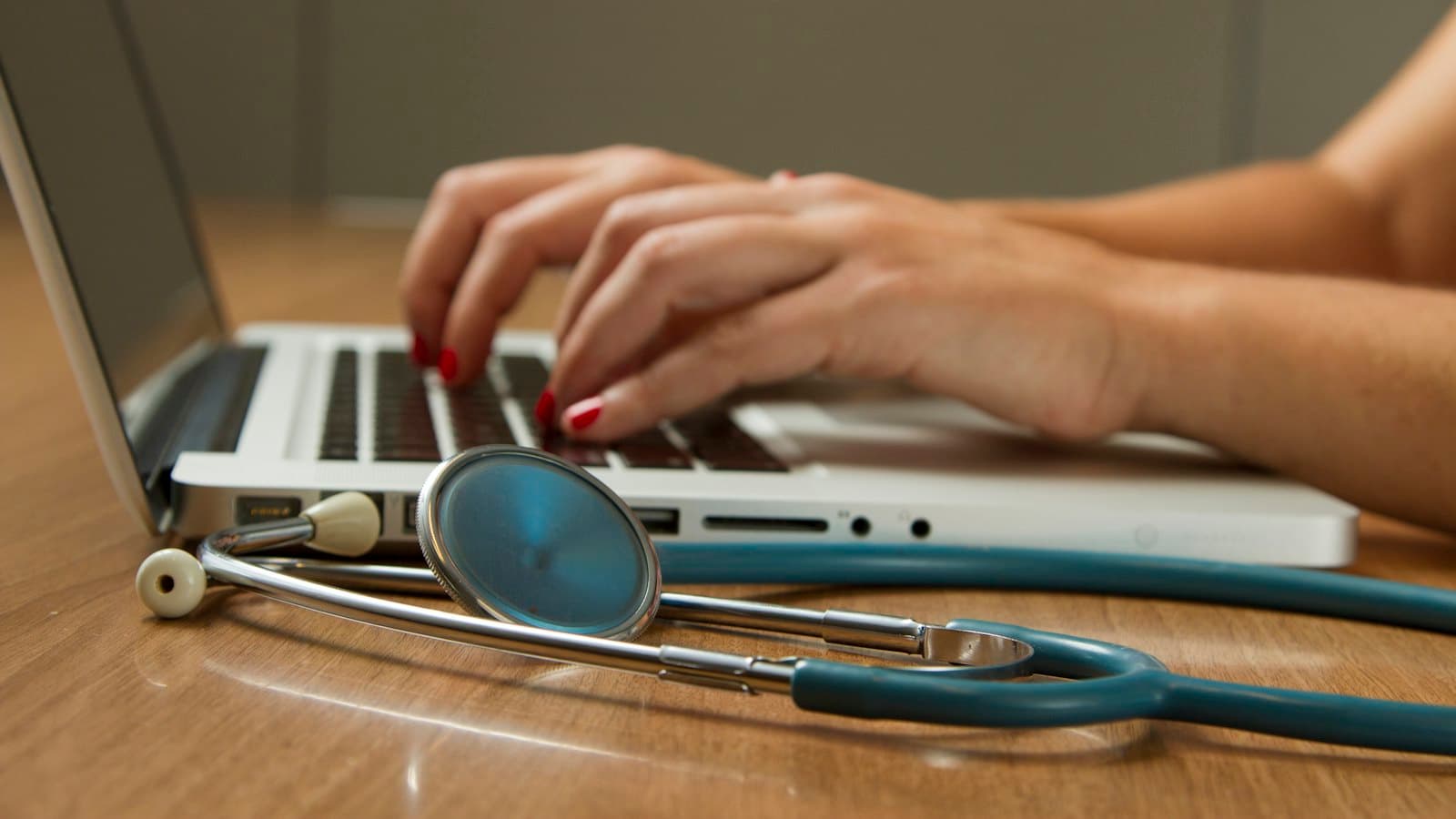 Hands typing on a laptop beside a stethoscope resting on a wooden desk