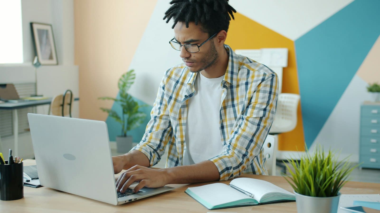 A young man in glasses typing on a laptop at a bright desk