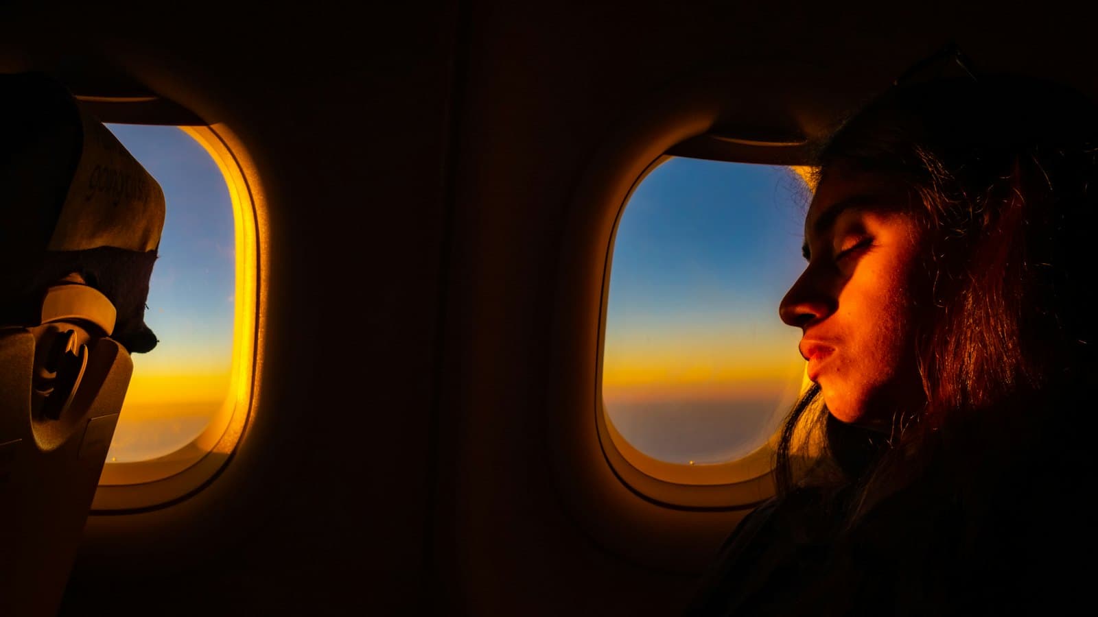 A woman resting her eyes beside an airplane window at golden sunset
