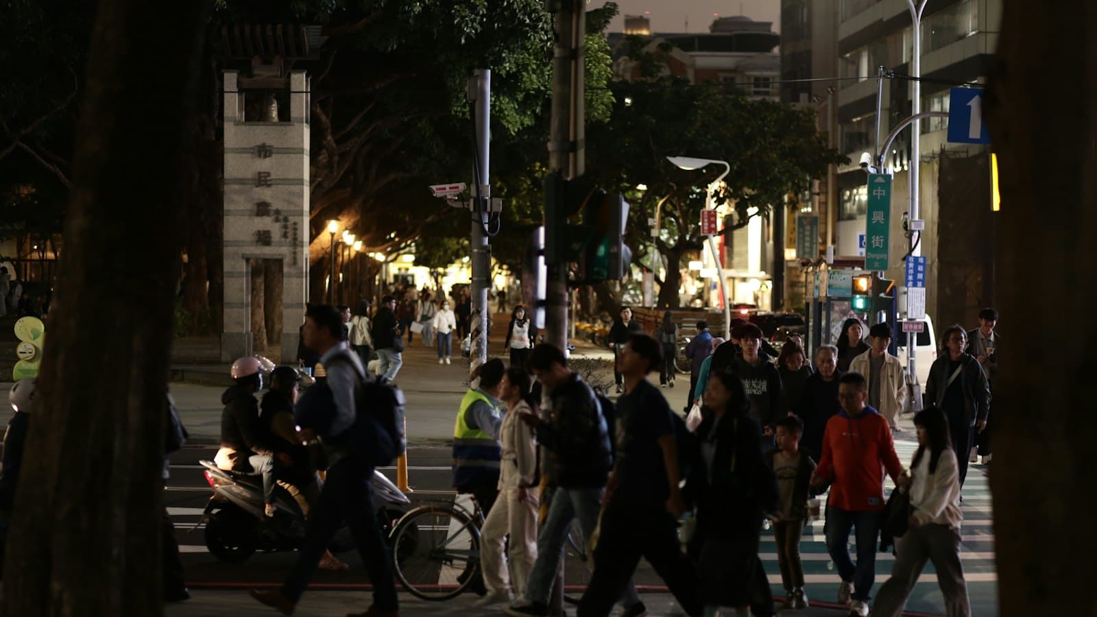 Evening crowds of pedestrians and families crossing a tree-lined street in Taipei