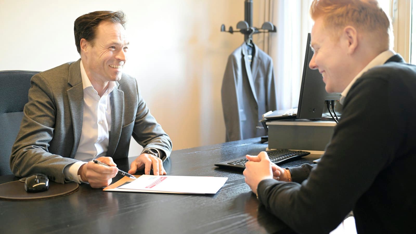 Two men smiling across an office desk while reviewing a document together