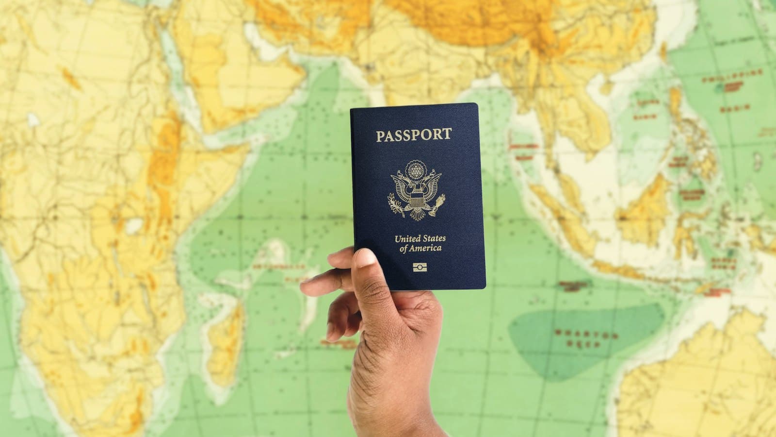 Hand holding up a passport in front of a colourful world map