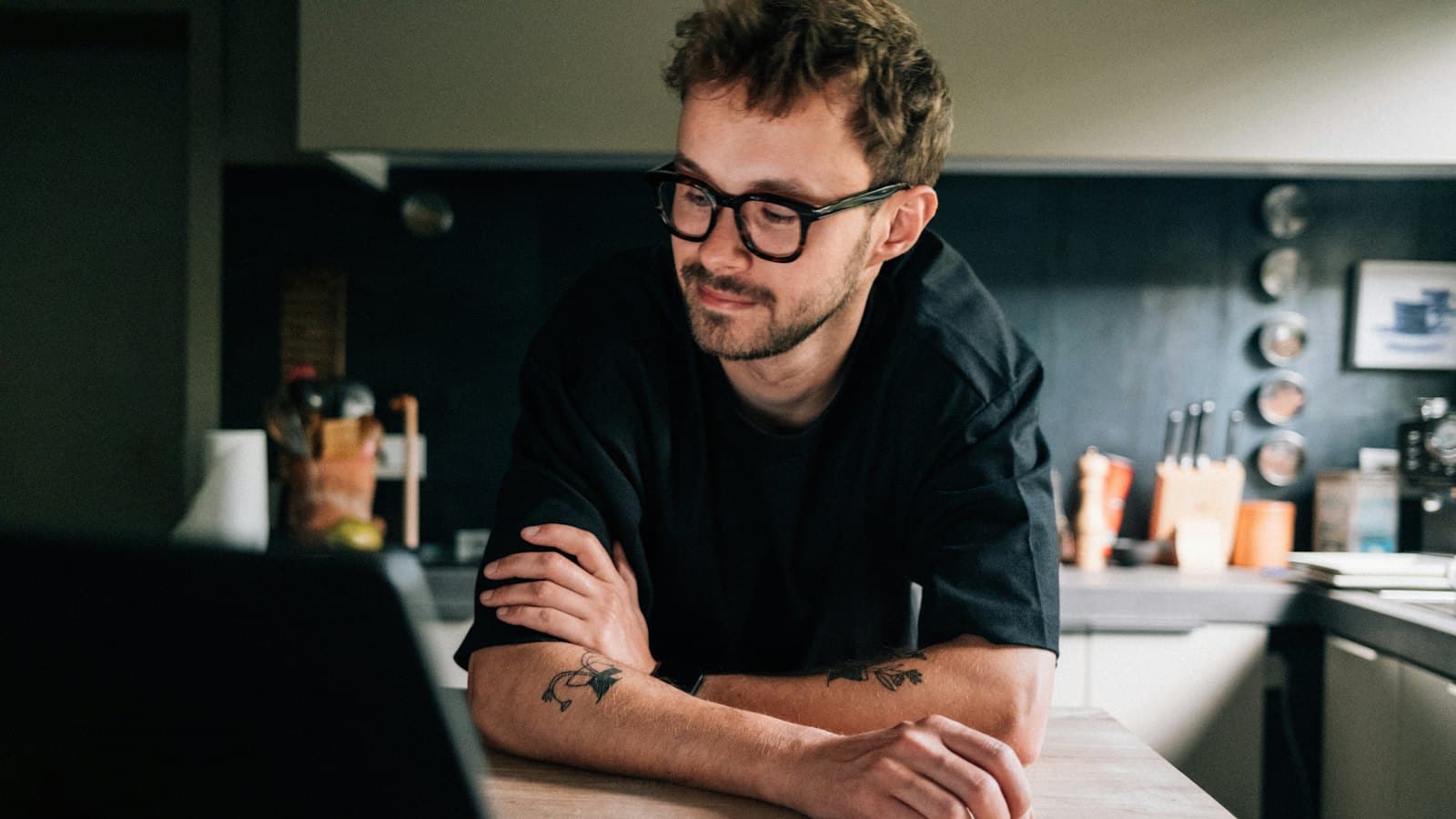 A man leaning on a kitchen counter, looking thoughtfully at his laptop