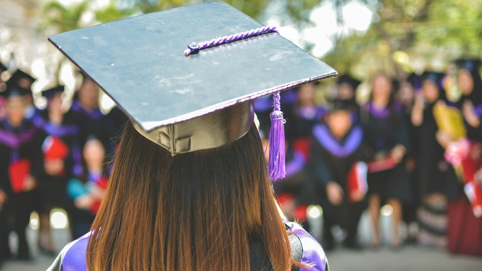 A graduate in cap and gown seen from behind, facing classmates at a ceremony
