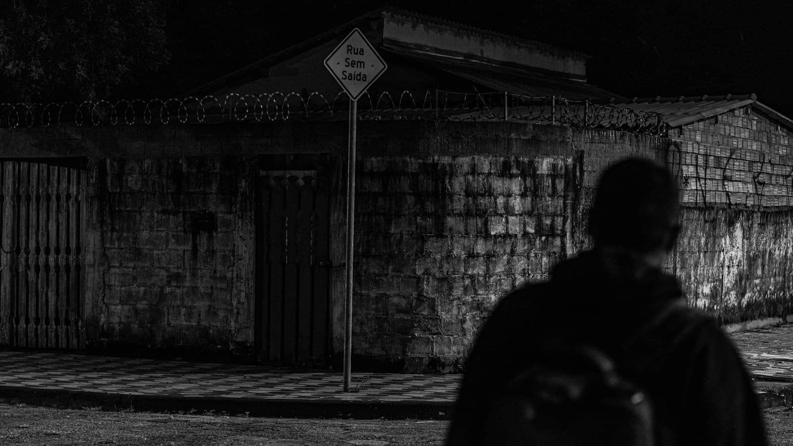 Black-and-white night photo of a person seen from behind, walking towards a walled building topped with razor wire beside a dead-end street sign