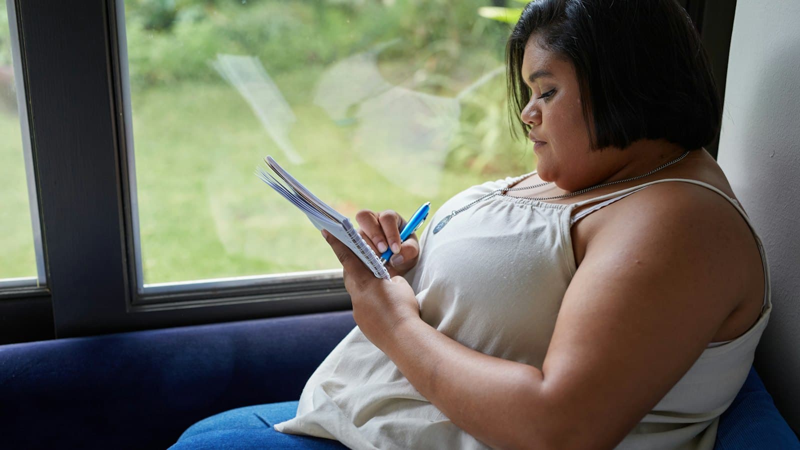 A woman sits on a blue couch by a window writing in a small notebook