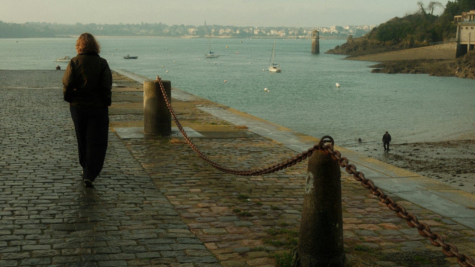 Woman walking along a cobblestone harbour path beside calm water and moored sailboats