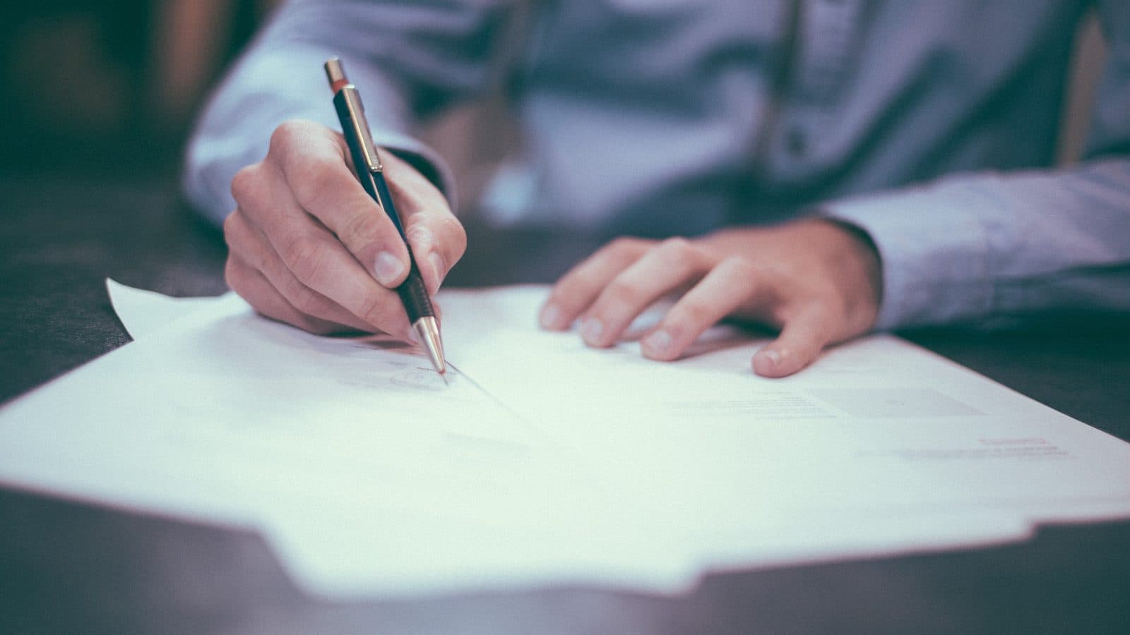 A man in a light blue shirt signing a paper document with a pen at a desk, face out of frame