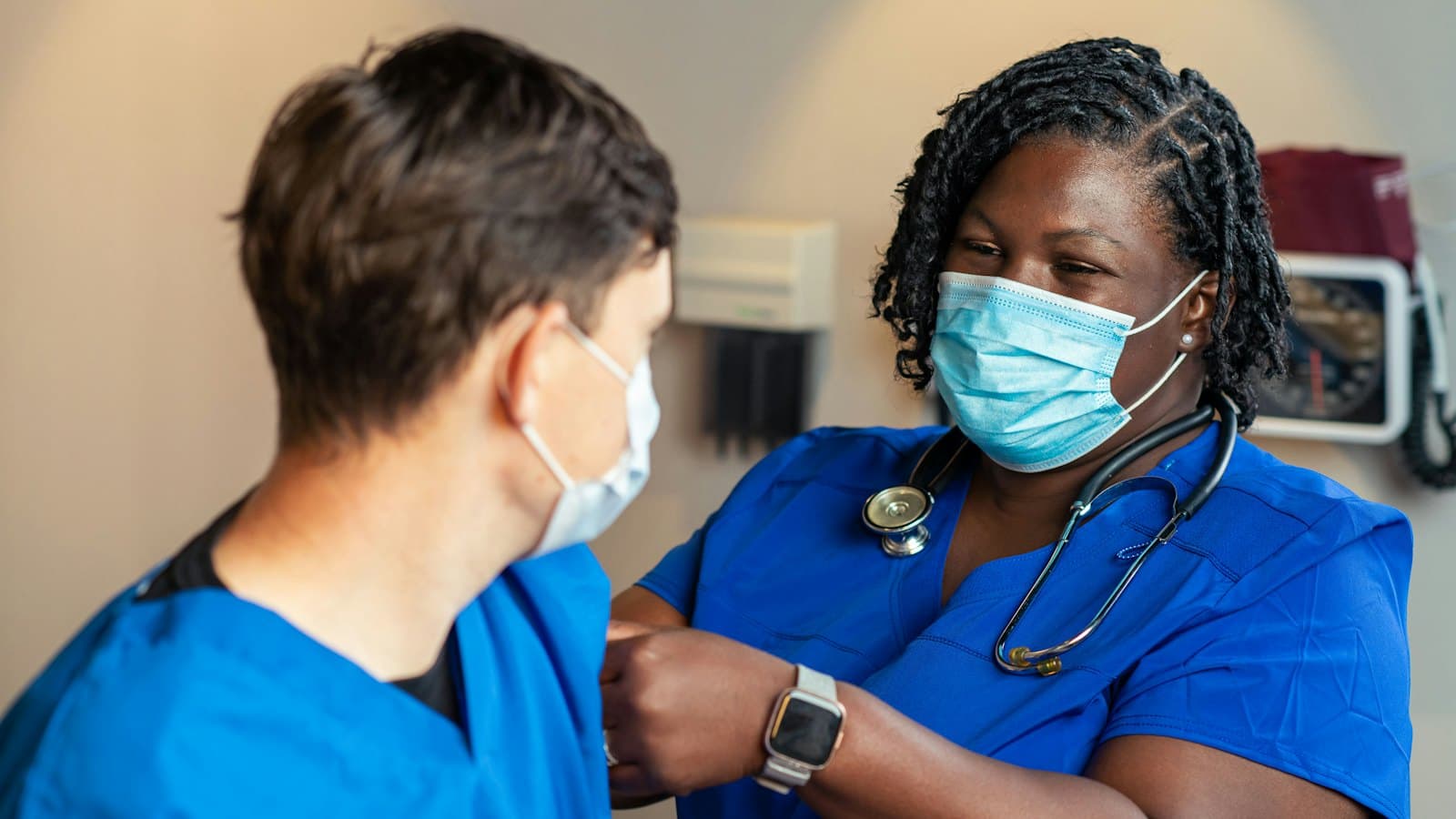 A nurse in blue scrubs and a stethoscope attending to a masked colleague in an exam room
