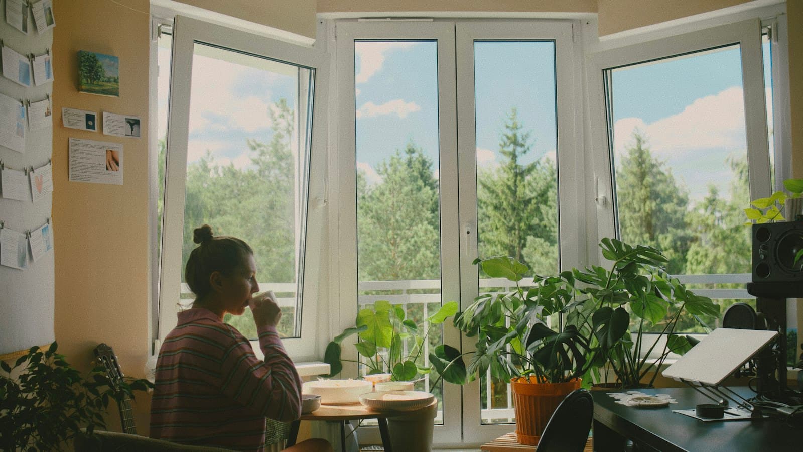 A woman sits quietly at a table by a sunlit window, holding a mug