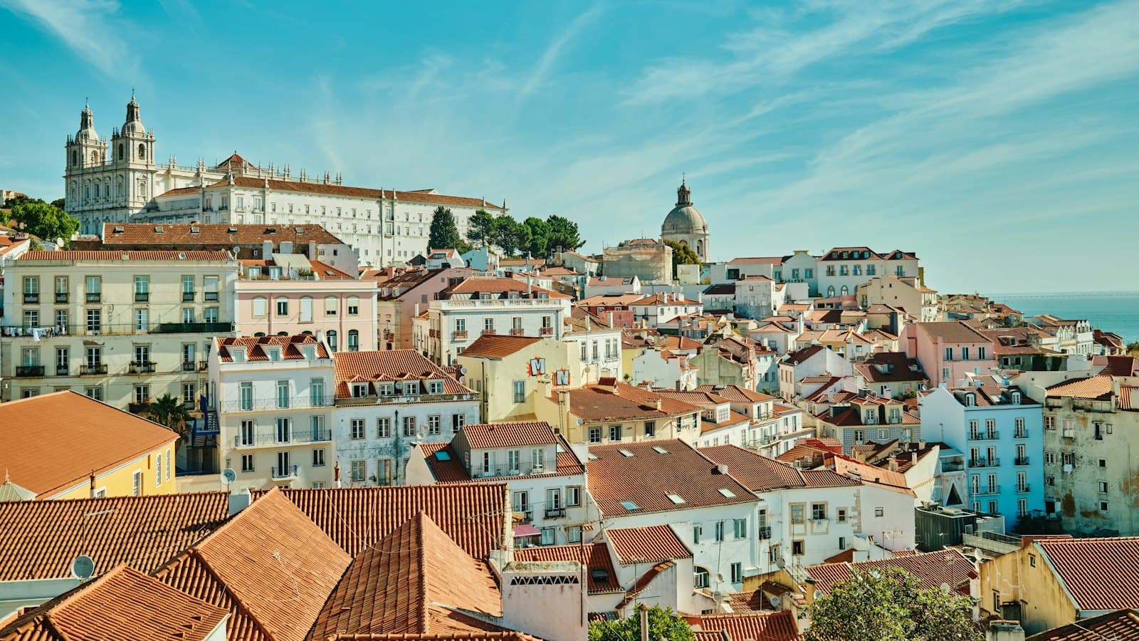 Terracotta rooftops of Lisbon's Alfama district below a white hilltop monastery on a sunny day
