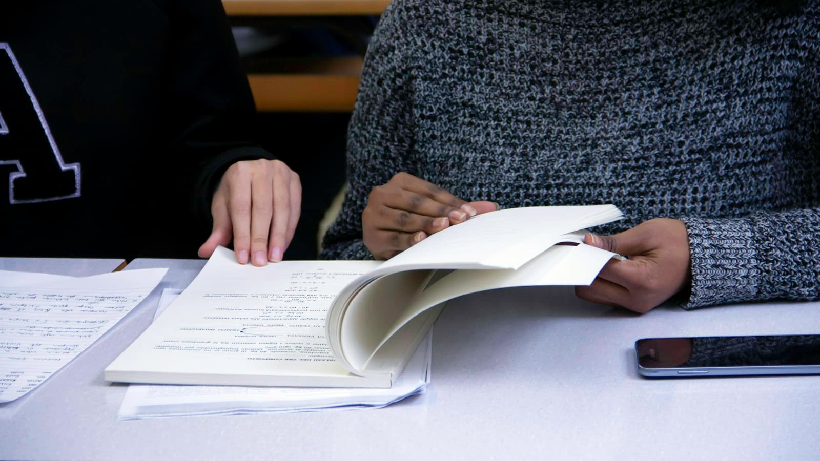 Two students turning through study papers at a classroom desk