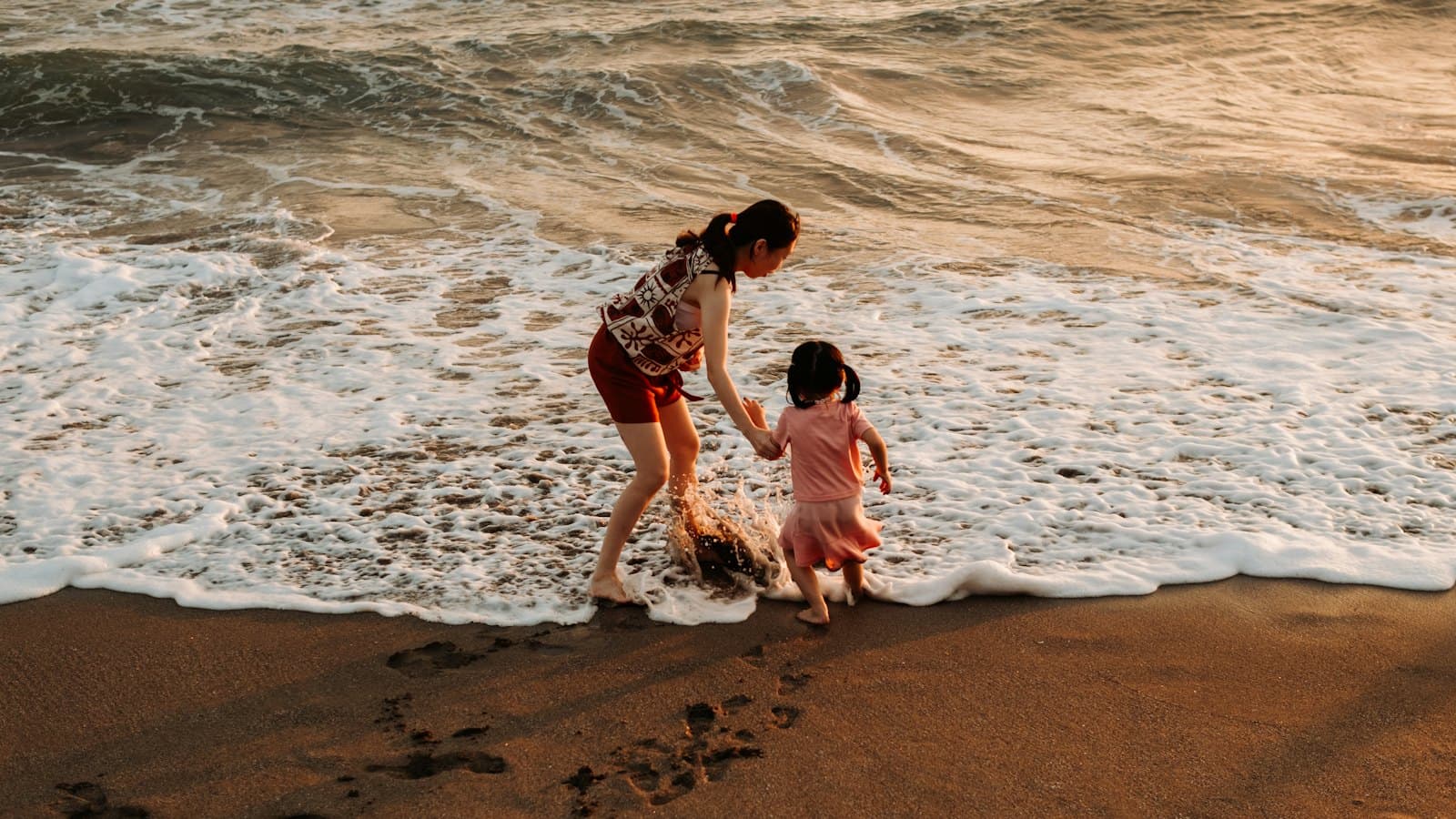 A mother and young daughter holding hands at the edge of the sea at sunset