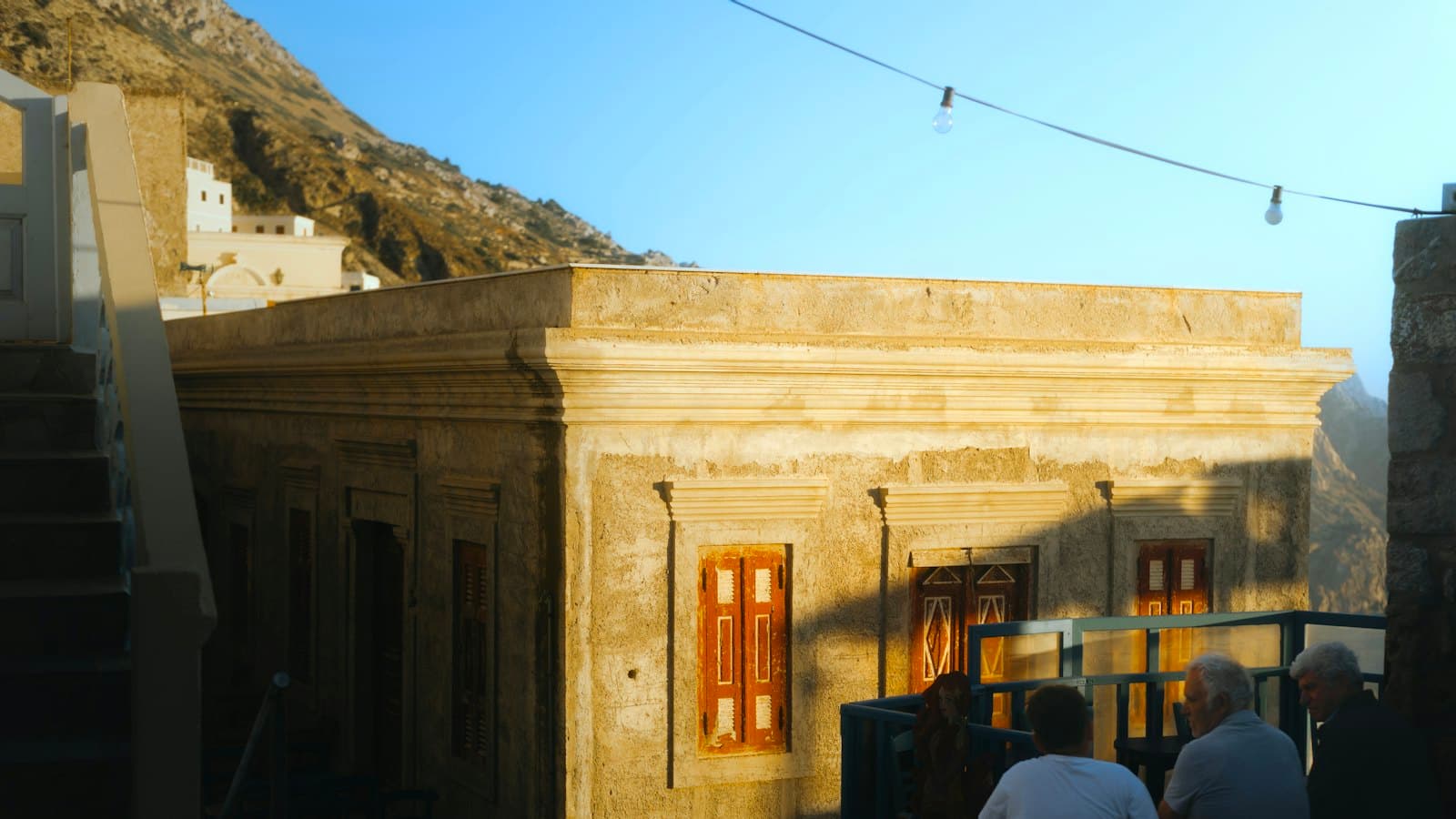 Older men chatting on a terrace beside a sunlit stone house in a Greek village