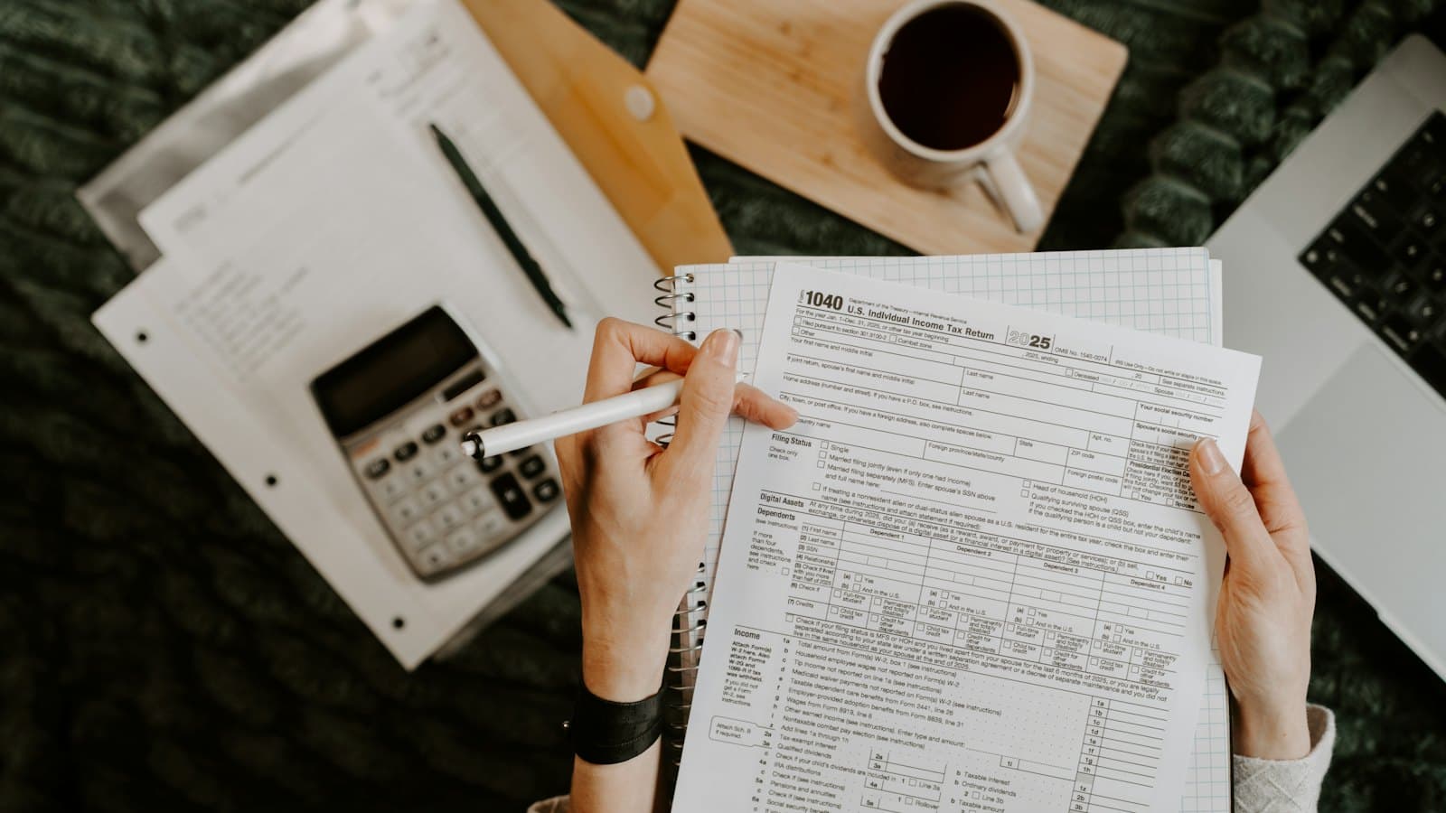 Hands holding a tax return form beside a calculator, coffee and laptop