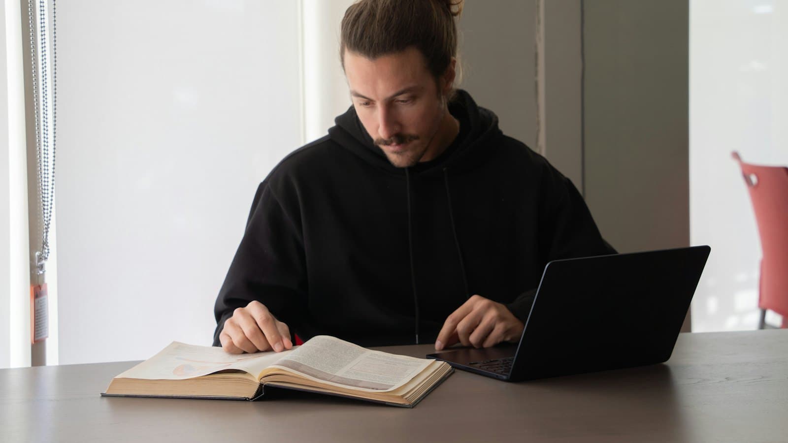 A man reading a book while working on his laptop at a desk