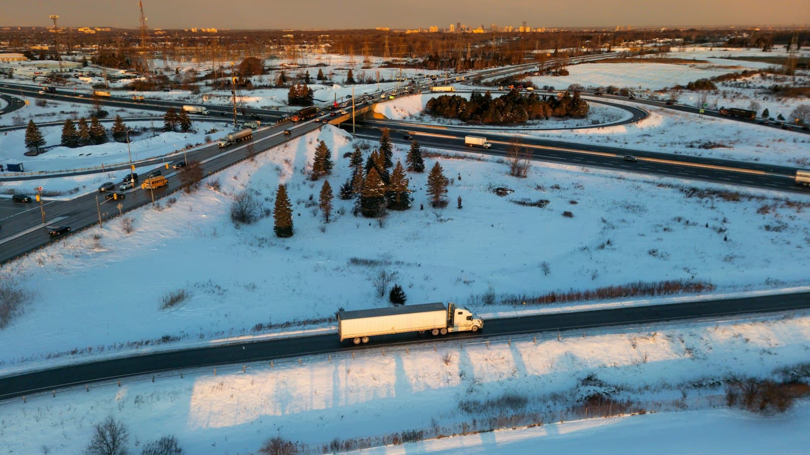 A white semi truck on a snow-covered highway near an interchange at sunset