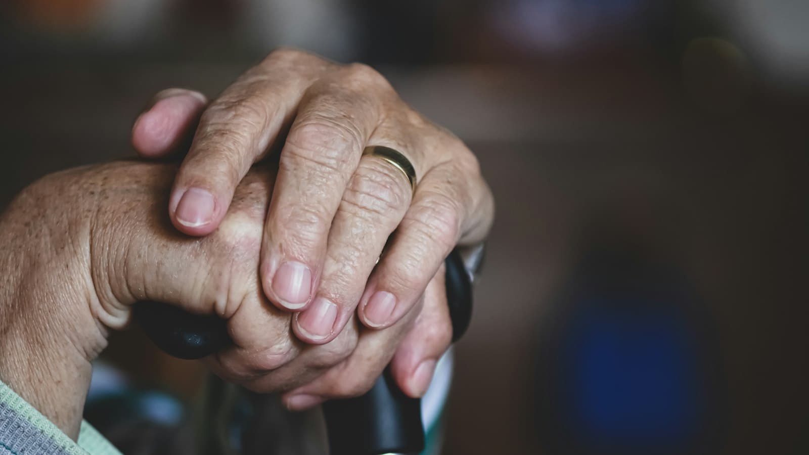 Close-up of an older person's wrinkled hands, wearing a gold ring, resting on a walking cane