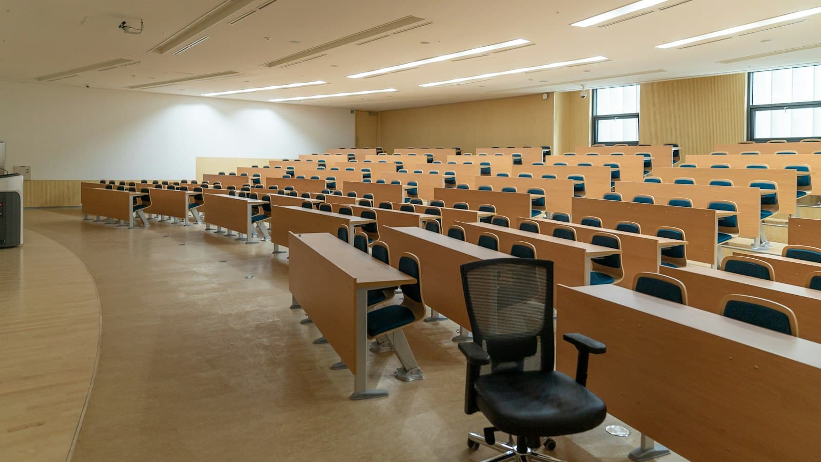 Rows of empty wooden desks with blue chairs in a large, silent university lecture hall